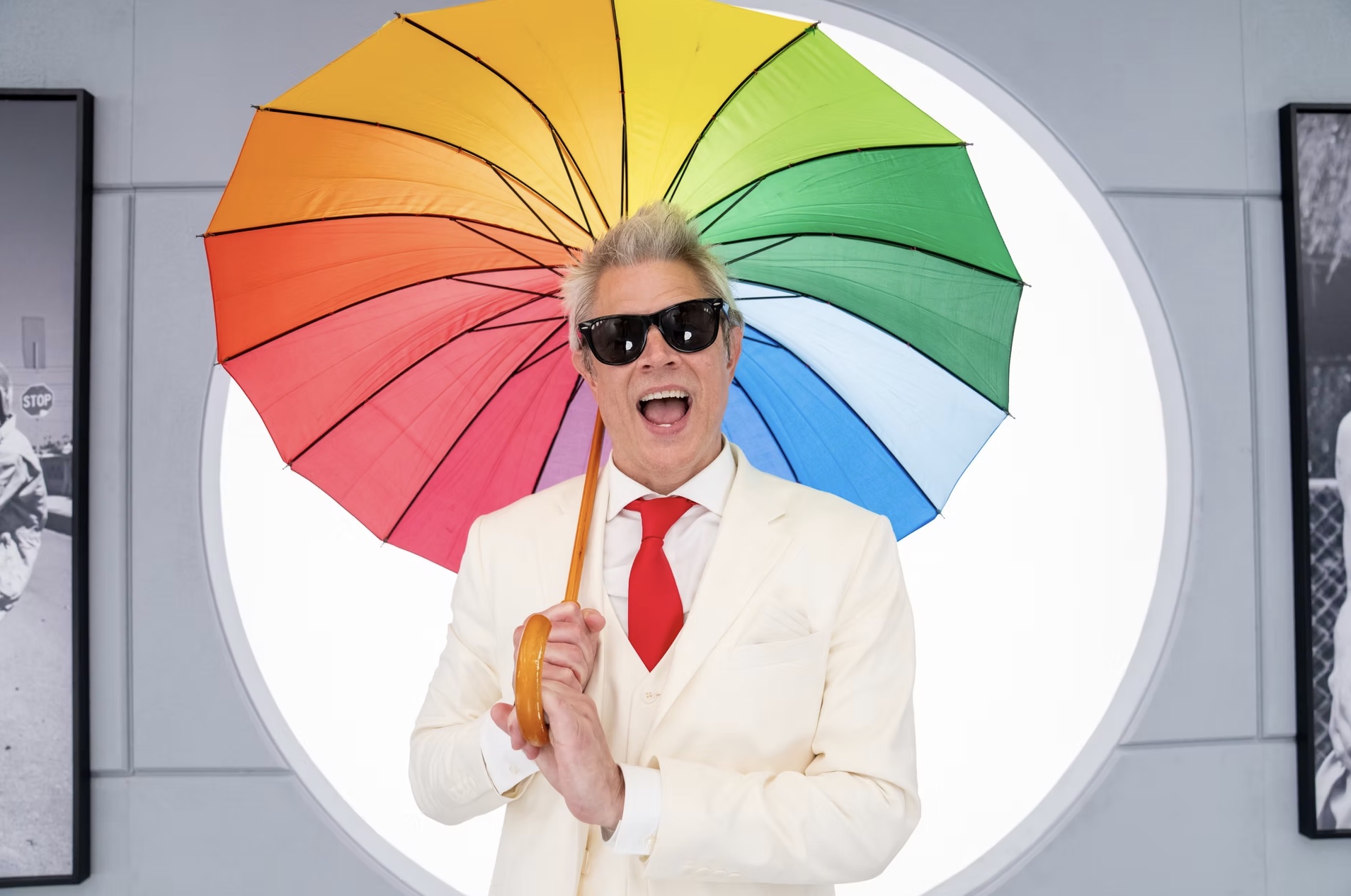 Johnny Knoxville holding a rainbow umbrella in front of a circular white background